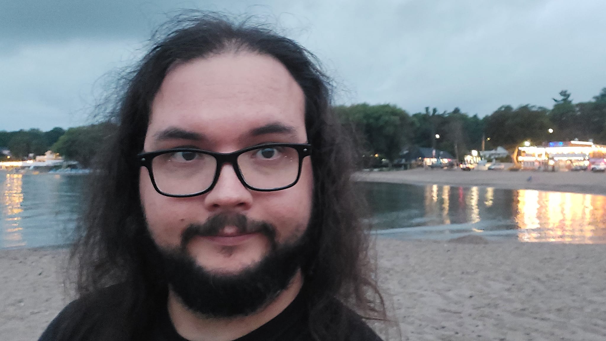 A man with long dark hair and a beard is at a sandy beach with a granite boulder breakwater, photo 7